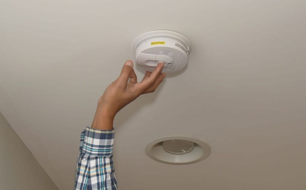 A hand reaching up to press a button on a smoke alarm that is on they ceiling.